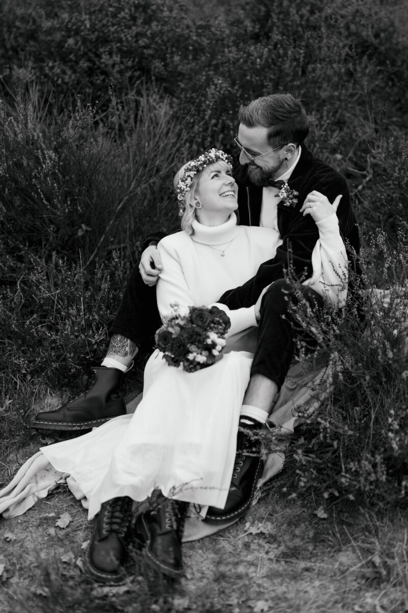 After Wedding, Braut und Bräutigam sitzen auf einer Wiese, sie liegt in seinen Armen. Beide lächeln sich an. Sie hält sich an seinem Oberarm fest. Bild ist in schwarz weiss.