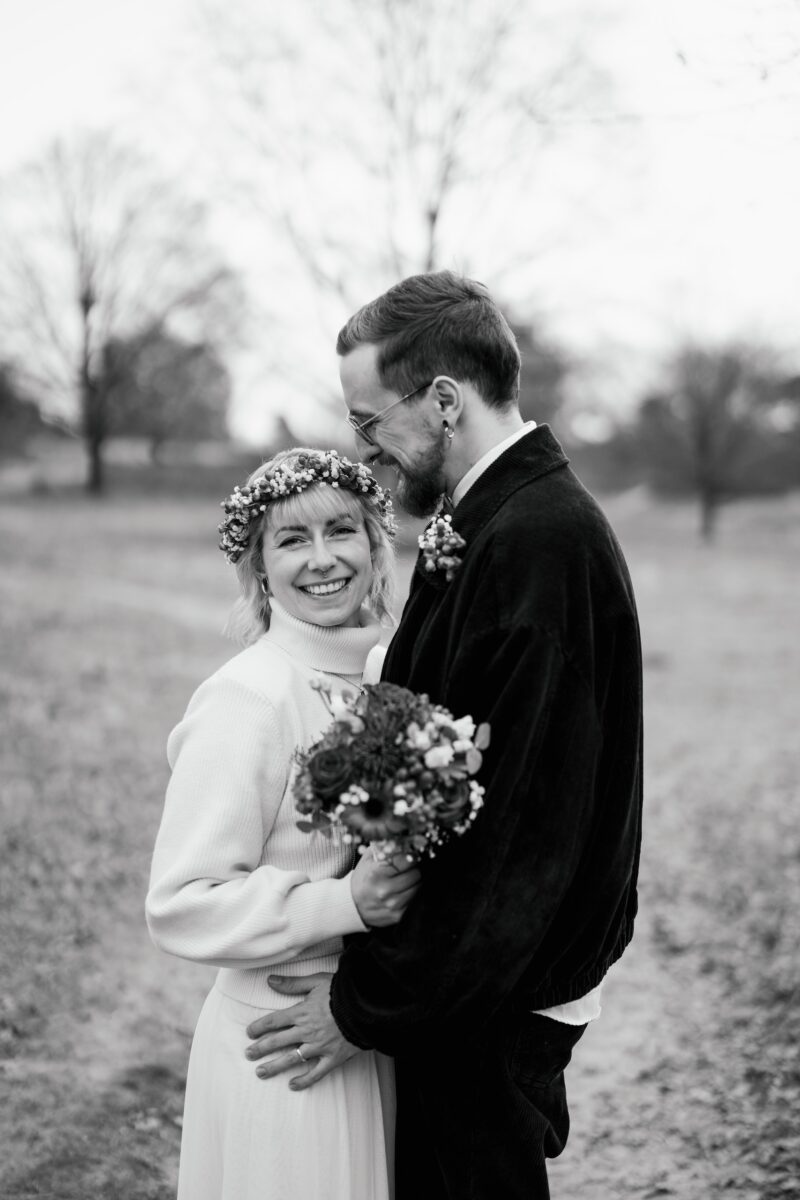 After Wedding, Braut und Bräutigam halten sich in den Armen. Er schaut sie verliebt an und lächelt sie an. Sie schaut lächelnd in die Kamera und hält einen Brautstrauß.