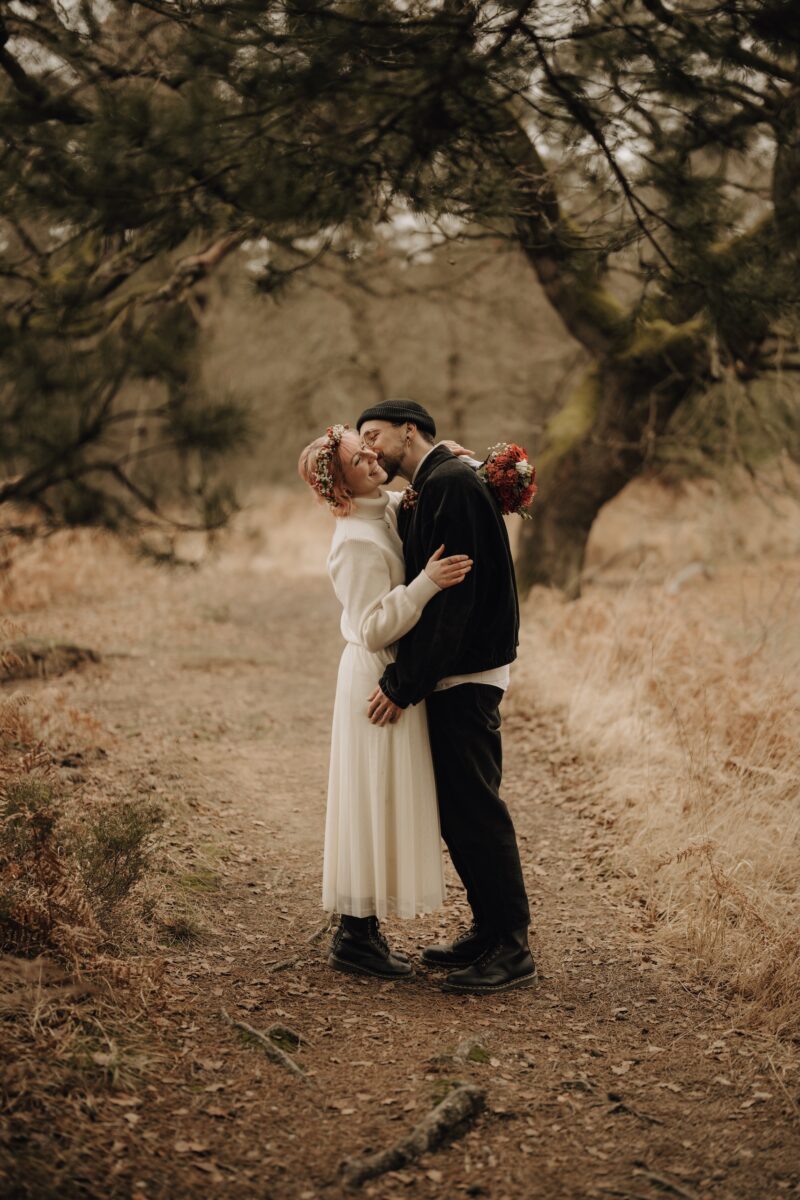 After Wedding Portfolio, Braut und Bräutigam stehen auf einer Lichtung. Beide umarmen sich und der Bräutigam küsst seine Braut auf die linke Wange, während sie lächelt