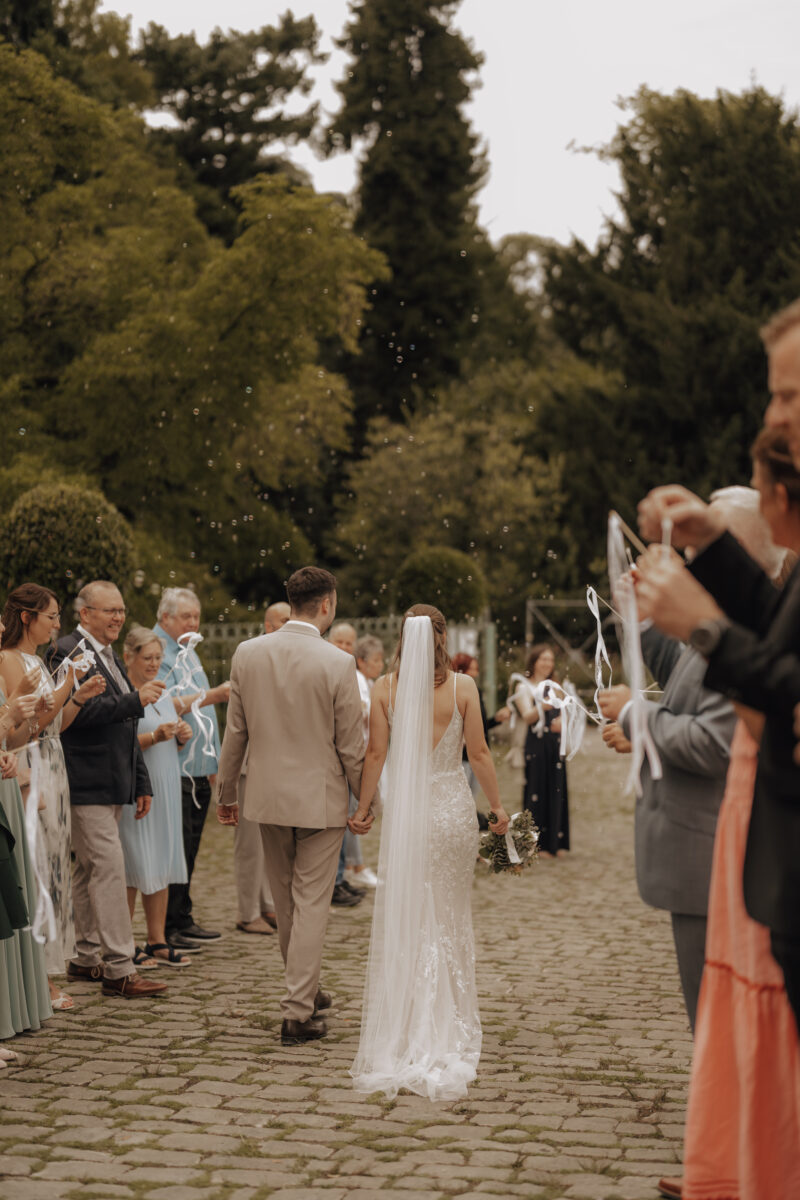 Hochzeitsportfolio, Hochzeitspaar läuft durch einen Spalier von ihren Gästen, die Gäste pusten Seifenblasen, das Brautpaar ist von hinten zu sehen