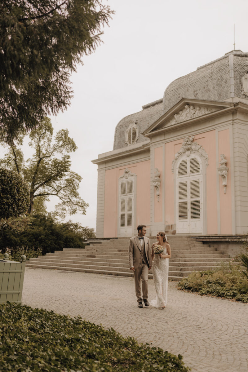 Hochzeitsportfolio, Brautpaar kommt Arm in Arm auf die Kamera zu, dabei gucken sie sich tief in die Augen, im Hintergrund sieht man einen Teil von einem Schloss