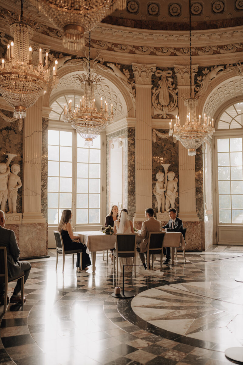 Hochzeitsportfolio, Aufnahme von hinten, zusehen ist der Trautisch im Schloss an dem das Hochzeitspaar, die Trauzeugen und die Standesbeamtin sitzt