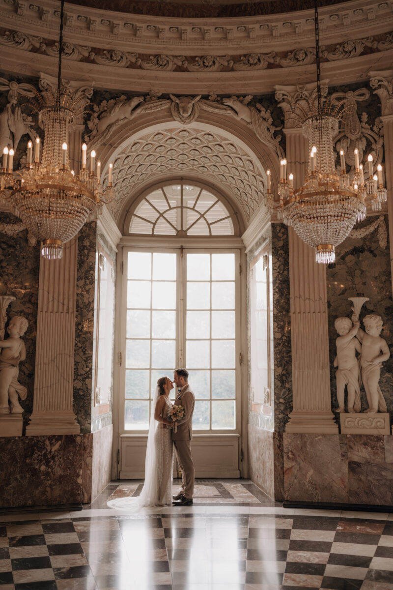 Hochzeitsportfolio, Brautpaar in prunkvollem Schloss mit Kronleuchtern, Brautpaar steht vor einem großen fenster gegenüber voneinander und schauen sich gegenseitig an | Hochzeitsfotografie NRW