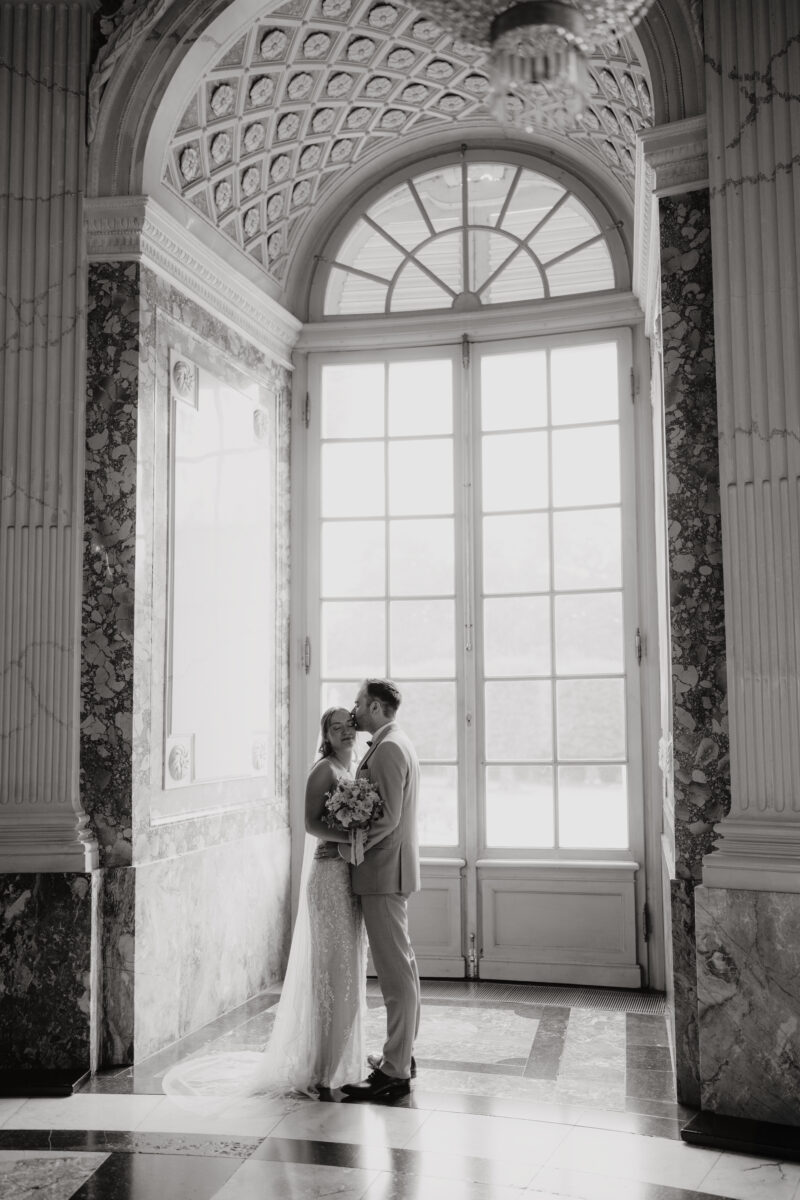 Hochzeitsportfolio, Brautpaar im eleganten Schlossfenster bei romantischem Hochzeitsshooting, Bräutigam nimmt Braut in den Arm und gibt ihr einen Kuss auf die Schläfe | Hochzeitsfotografin Köln