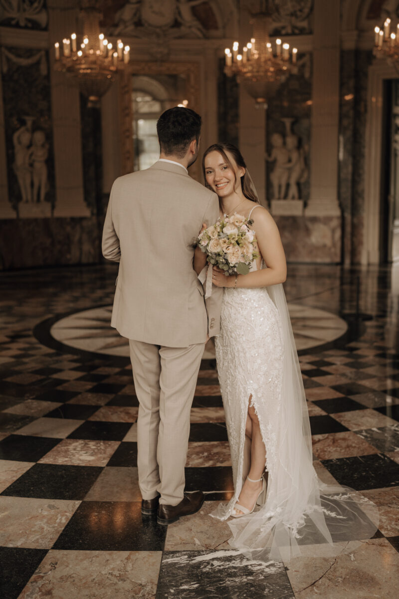 Hochzeitsportfolio, Brautpaar im eleganten Schlossinneren, Bräutigam steht mit dem Rücken zum Fotografen, Braut lehnt sich an seinen Arm und lächelt in die Kamera | Hochzeitsfotografie Köln