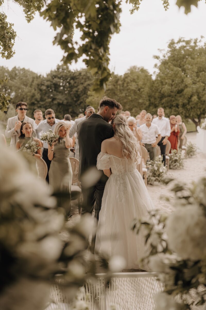 Hochzeitsportfolio, Braut und Bräutigam vom Altar nach draußen. Sie ist bei ihm eingehackt und hat ihren Kopf an seine Schulter gelegt. Im Hintergrund die Hochzeitsgäste.