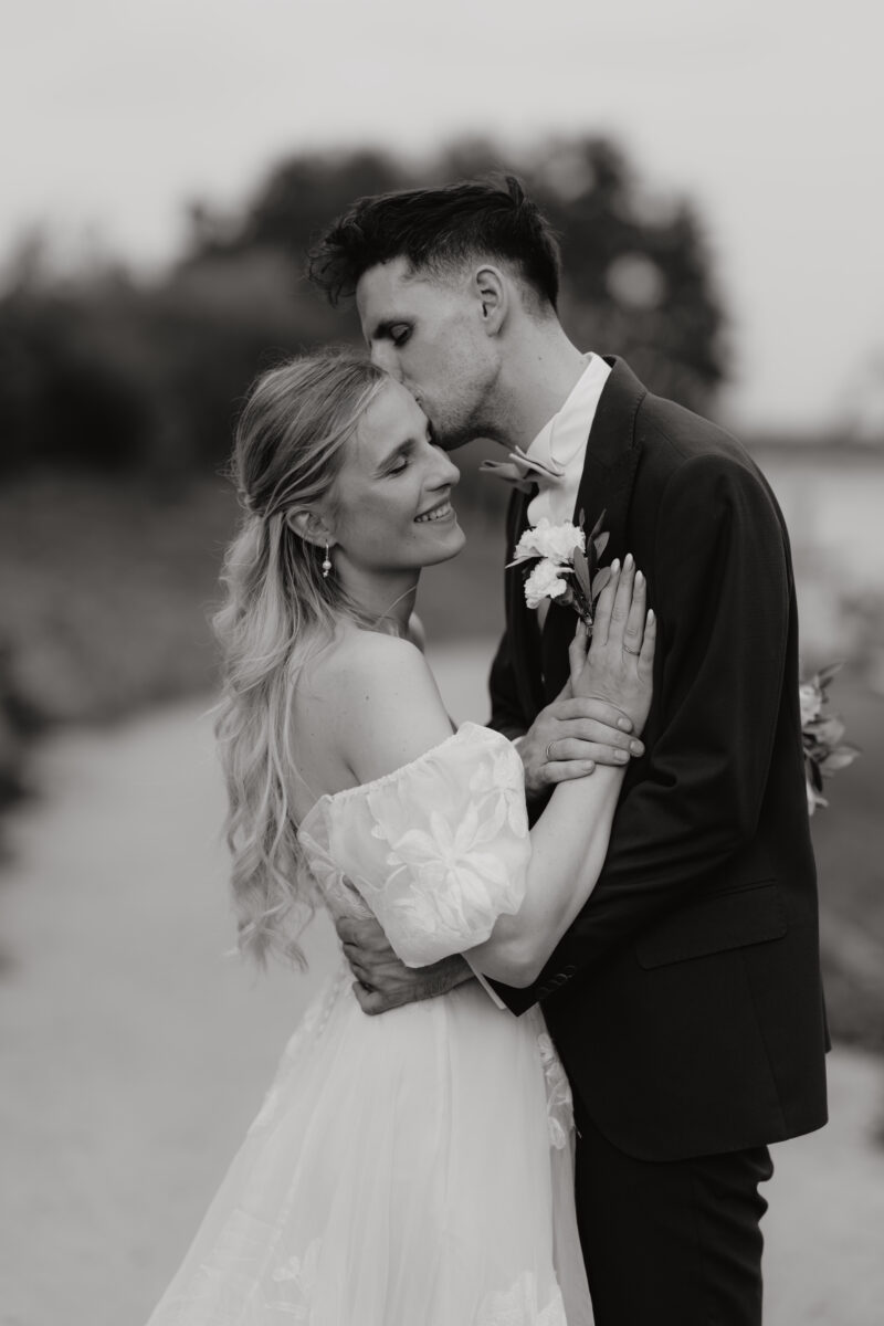 Hochzeitsportfolio, Braut und Bräutigam halten sich fest. Die Hand der Braut liegt auf seiner Brust. Er gibt ihr einen Kuss auf die Schläfe