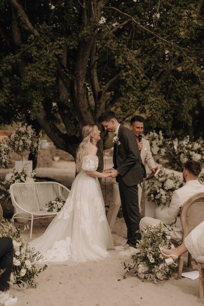 Hochzeitsportfolio, Braut und Bräutigam halten sich an den Händen. Bräutigam lehnt sich zu ihr. Sie lächeln sich beide an.