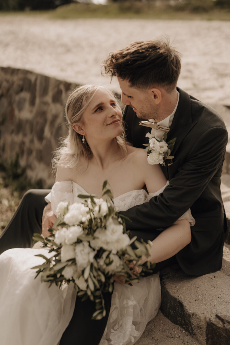 Hochzeitsportfolio, Braut und Bräutigam sitzen mit Brautstrauß in der Abendsonne, Bräutigam umarmt sie von hinten, sie schauen sich beide tief in die Augen