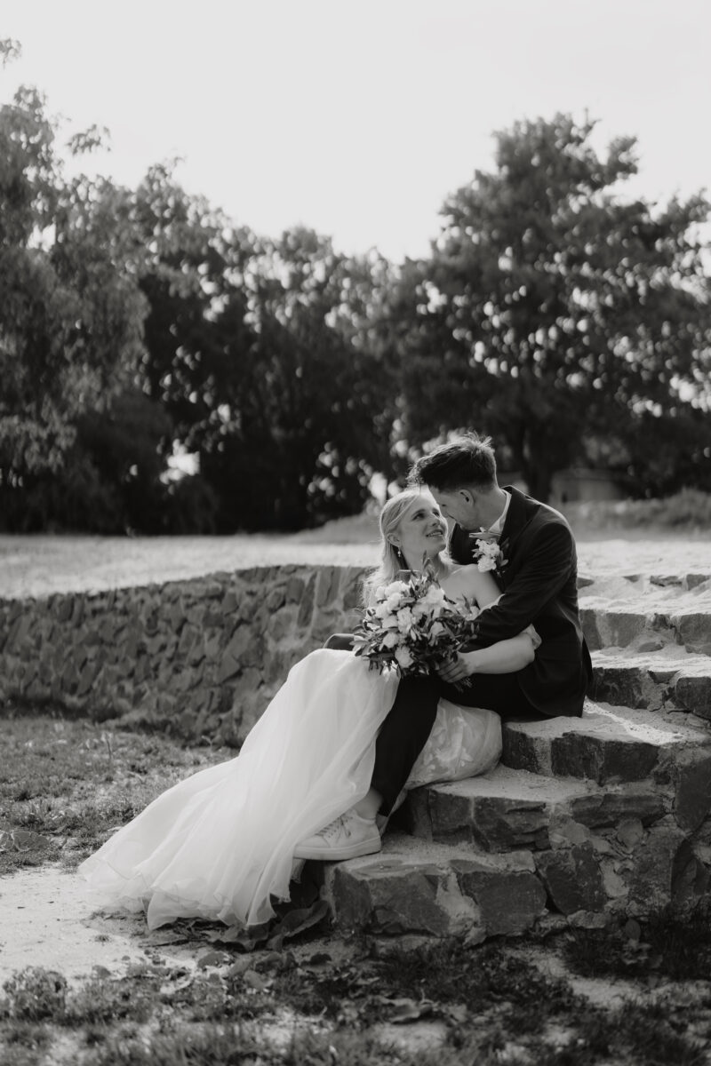 Hochzeitsportfolio, der Bräutigam sitzt auf einer kleinen Steintreppe. Die Braut liegt in seinen Armen und beide schauen sich lächelnd an. Die Braut hält ihren Brautstrauß in der Hand.