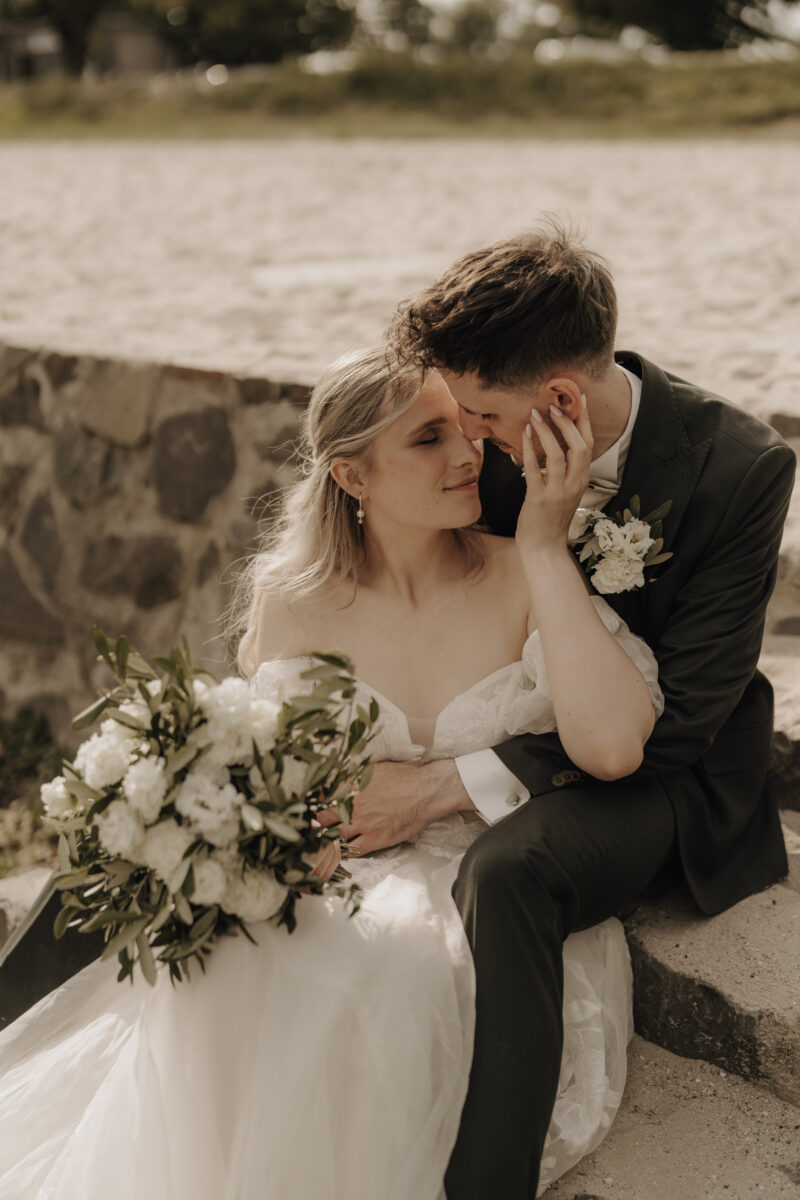 Hochzeitsportfolio, Braut und Bräutigam sitzen mit Brautstrauß in der Abendsonne, Bräutigam umarmt sie von hinten | Hochzeitsfotografie Köln