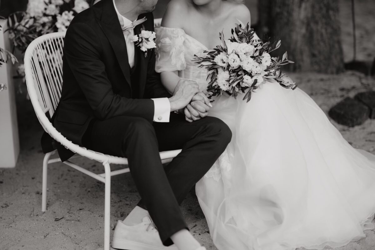 Hochzeitsportfolio, Braut und Bräutigam sitzen auf Stühlen nebeneinander. Der Bräutigam hält die Hand seiner Braut