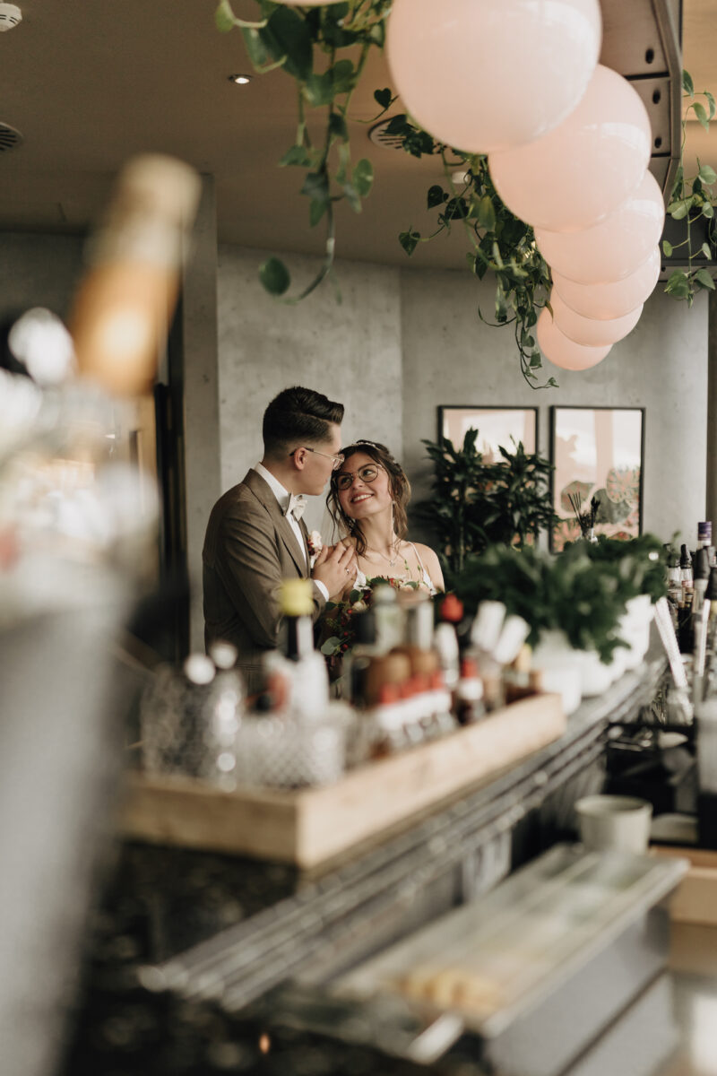 Hochzeitsportfolio, Braut und Bräutigam stehen hinter einer Cocktailtheke. Sie schaut lächelnd zu ihm hoch. Er hält sie an der Schulter.