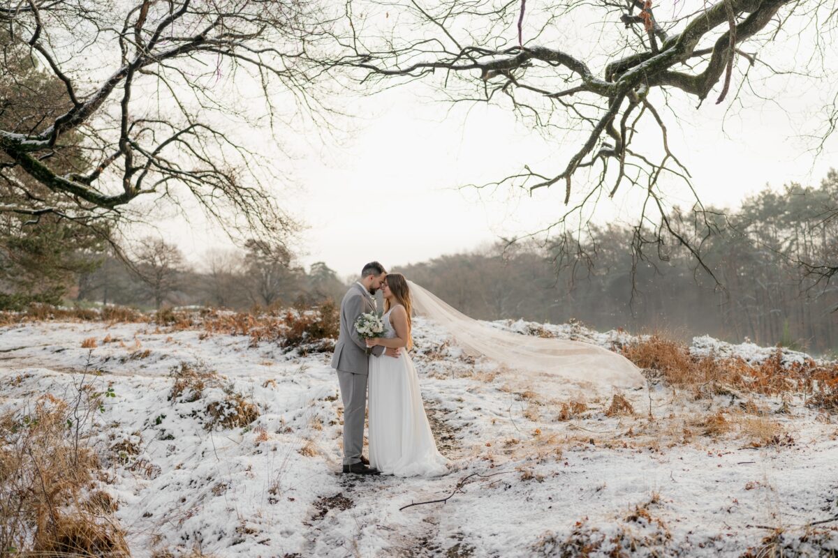 After Wedding Portfolio, Braut und Bräutigam stehen im Schnee, umgeben von Bäumen. Beide halten sich gegenseitig an der Taille. Ihre Stirnen liegen ganz nah aneinander. Der Schleier der Braut weht nach hinten.