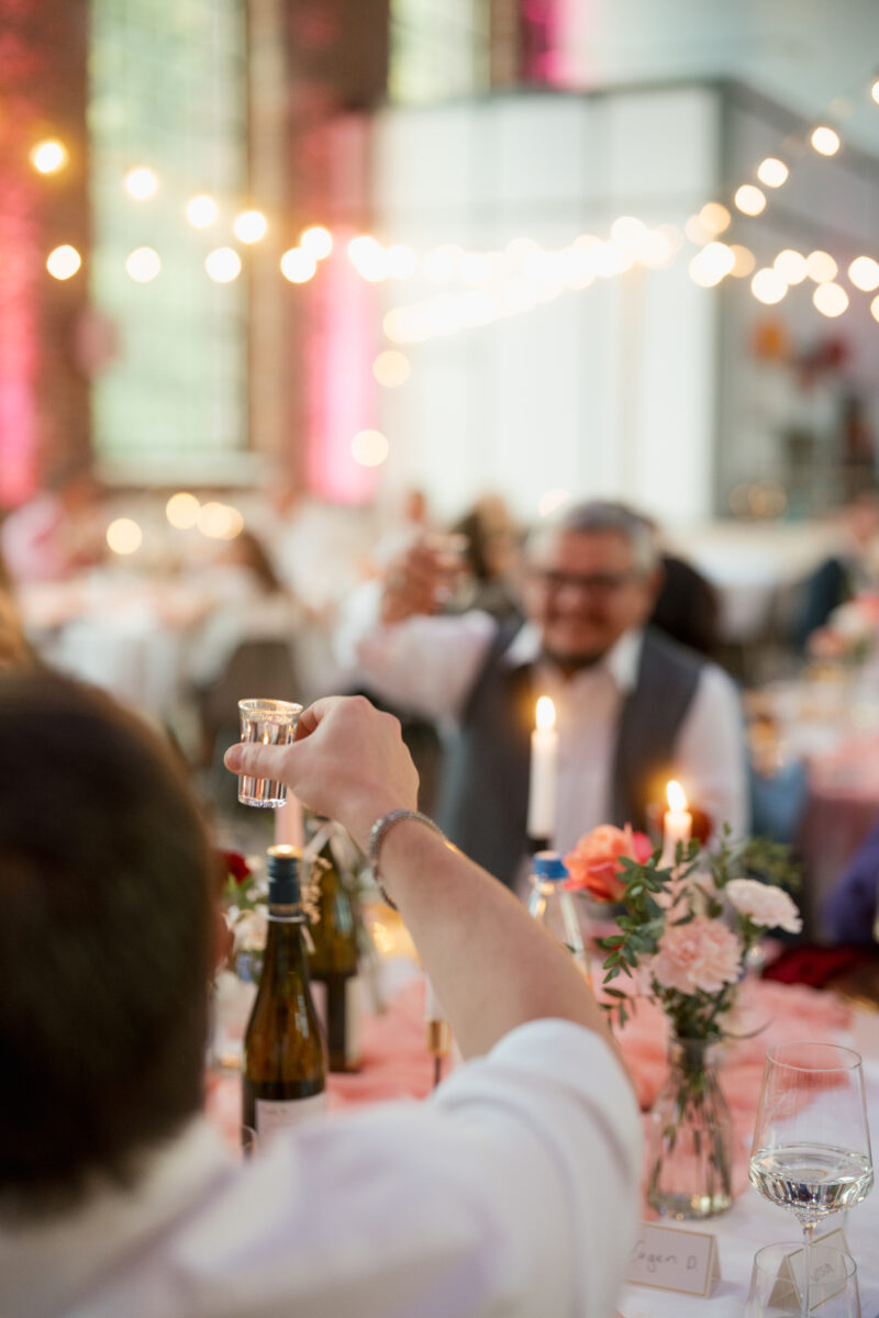 Hochzeitsportfolio, Wodka shot ist im Fokus, Detailaufnahme, Hochzeitsgäste stoßen gemeinsam an