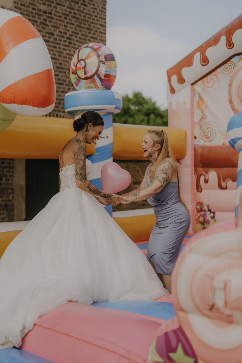 Hochzeitsportfolio, Braut und Freundin lachen gemeinsam auf einer Hüpfburg während der Hochzeitsfeier | Hochzeitsreportage Köln