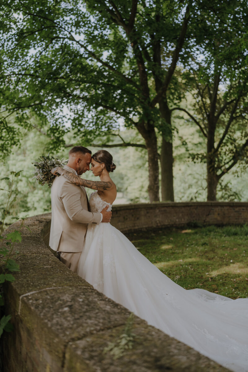 Hochzeitsportfolio, Brautpaarshooting unter einem Baum im weichen Licht, Bräutigam lehnt sich an eine Mauer, Braut steht im gegenüber, Stirn an Stirn und haben beide die Augen geschlossen | Hochzeitsfotografin Köln