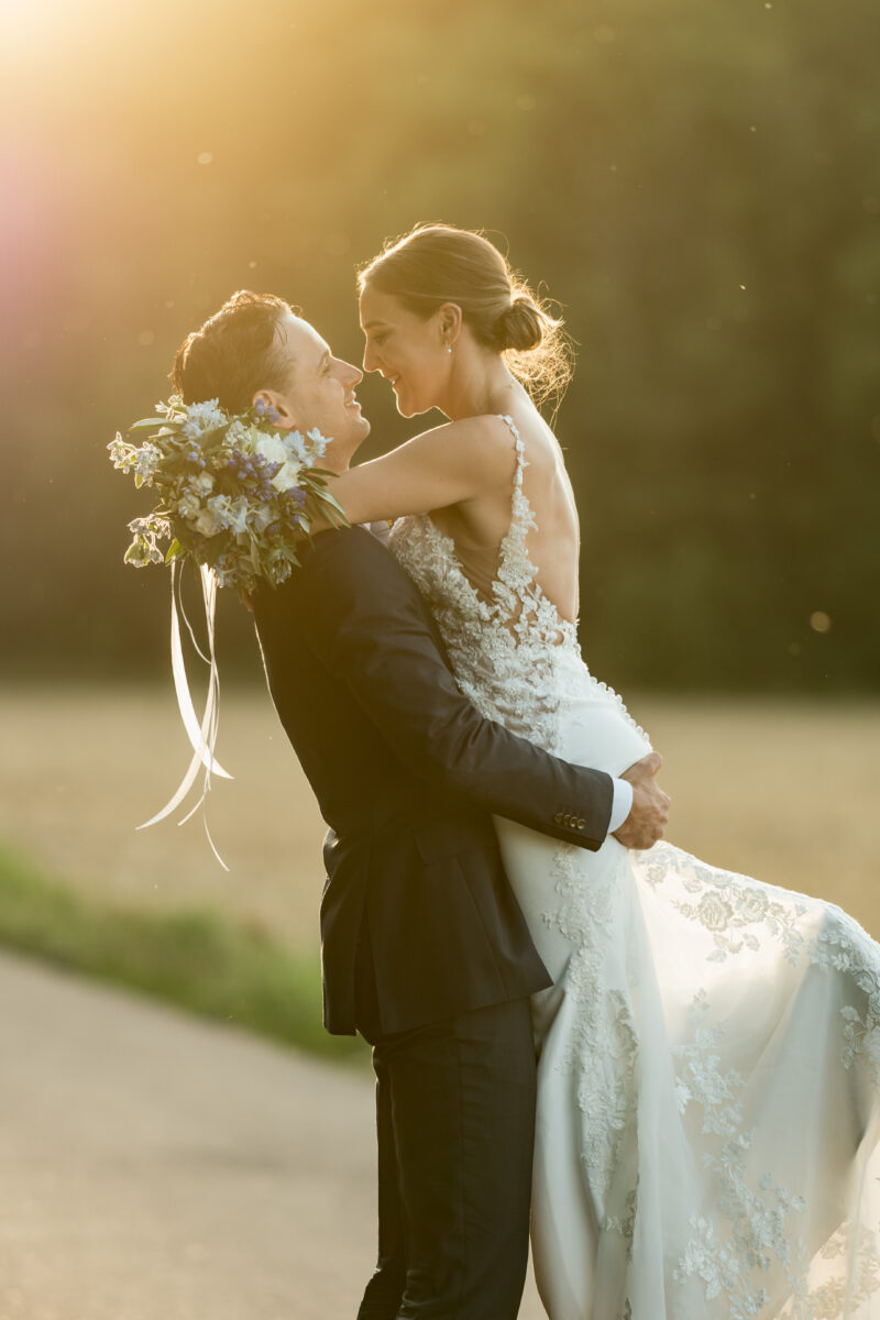 Hochzeitsportfolio, Hochzeitspaar wird in der Abendsonne fotografiert, Bräutigam hebt die Braut hoch und sie lächeln sich an