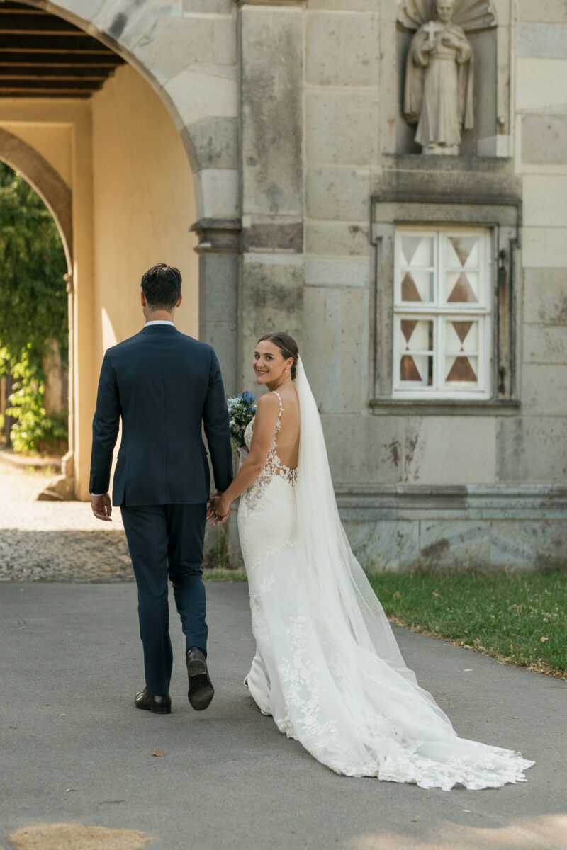 Hochzeitsportfolio, Brautpaar geht Hand in Hand vom Fotografen weg, dabei schaut die Braut in die Kamera