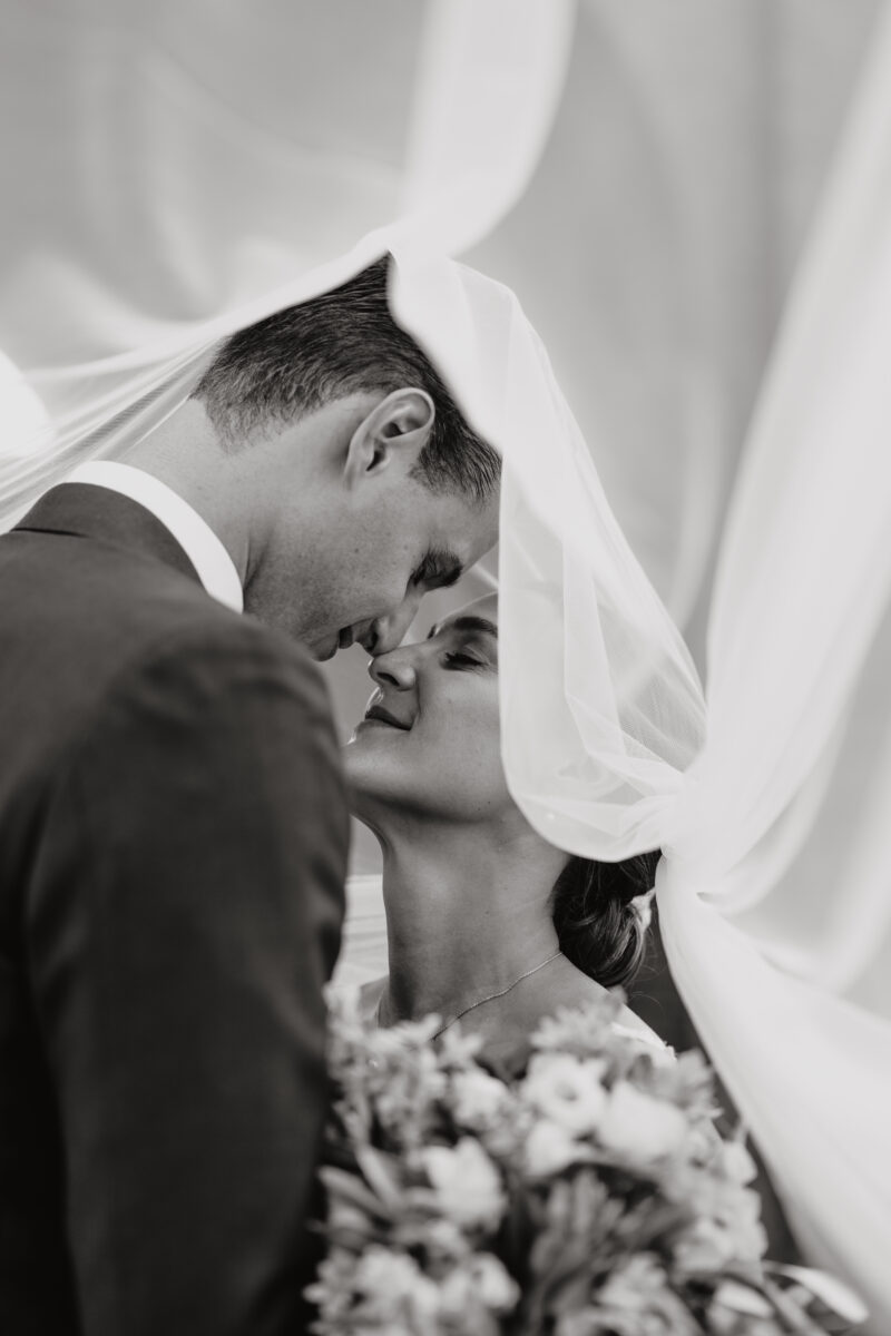 Hochzeitsportfolio, Hochzeitspaar ist vom Schleier der Braut umhüllt, stehen gegenüber voneinander, Nasenspitze an Nasenspitze und beide haben ihre Augen zu