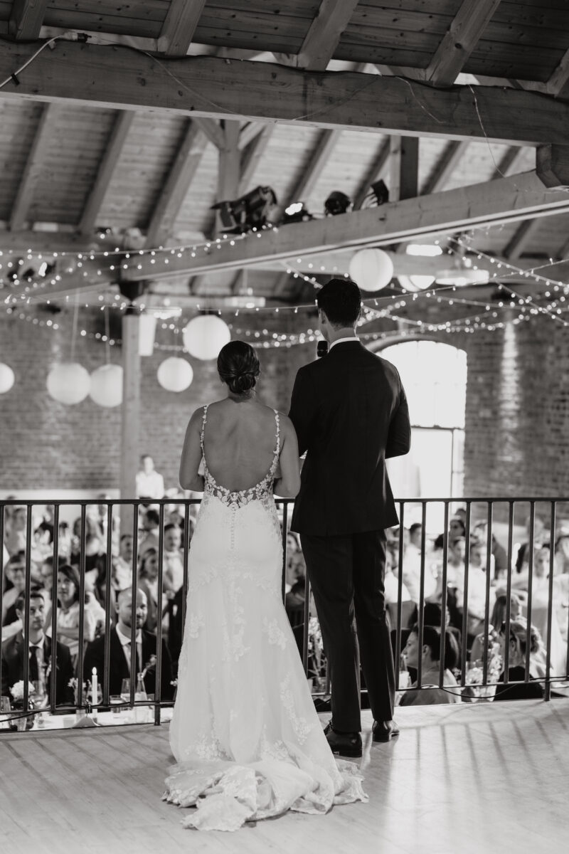 Hochzeitsportfolio, Das Brautpaar steht auf einer Art Podium zu ihren Gästen gewandt, das Paar steht mit dem Rücken zum Fotografen, es ist eine Schwarz-Weiß Aufnahme