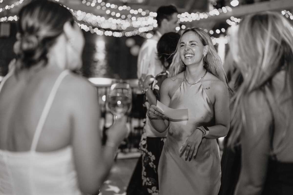 Hochzeitsportfolio, Freundin der Braut steht im Fokus und ist mit der Braut am lachen. Brautjunfer lacht herzlich die Braut an