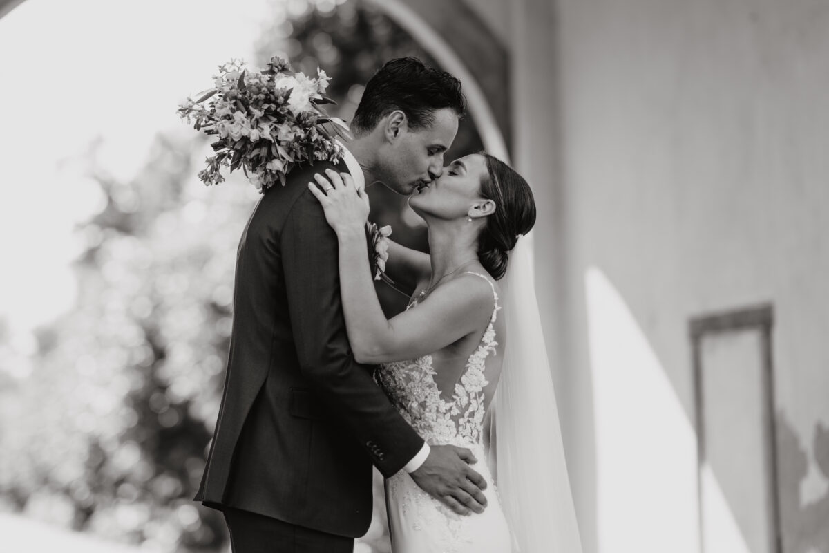 Hochzeitsportfolio, Hochzeitspaar steht gegenüber voneinander und küsst sich, sie stehen unter einem Torbogen, die Aufnahme ist in schwarz-weiß