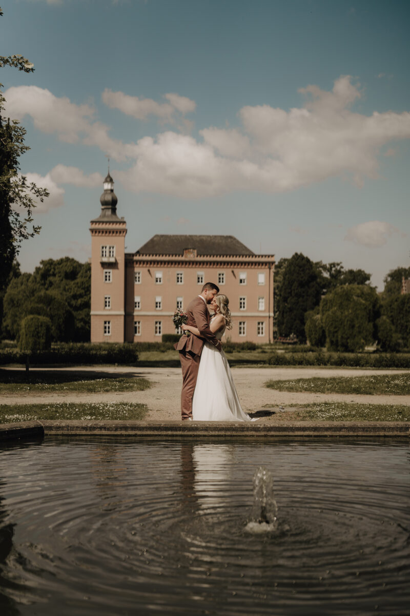 Hochzeitsportfolio, Braut und Bräutigam stehen vor einem Schloss im Vordergrund ist ein kleiner See. Sie halten sich in den Armen und küssen sich.
