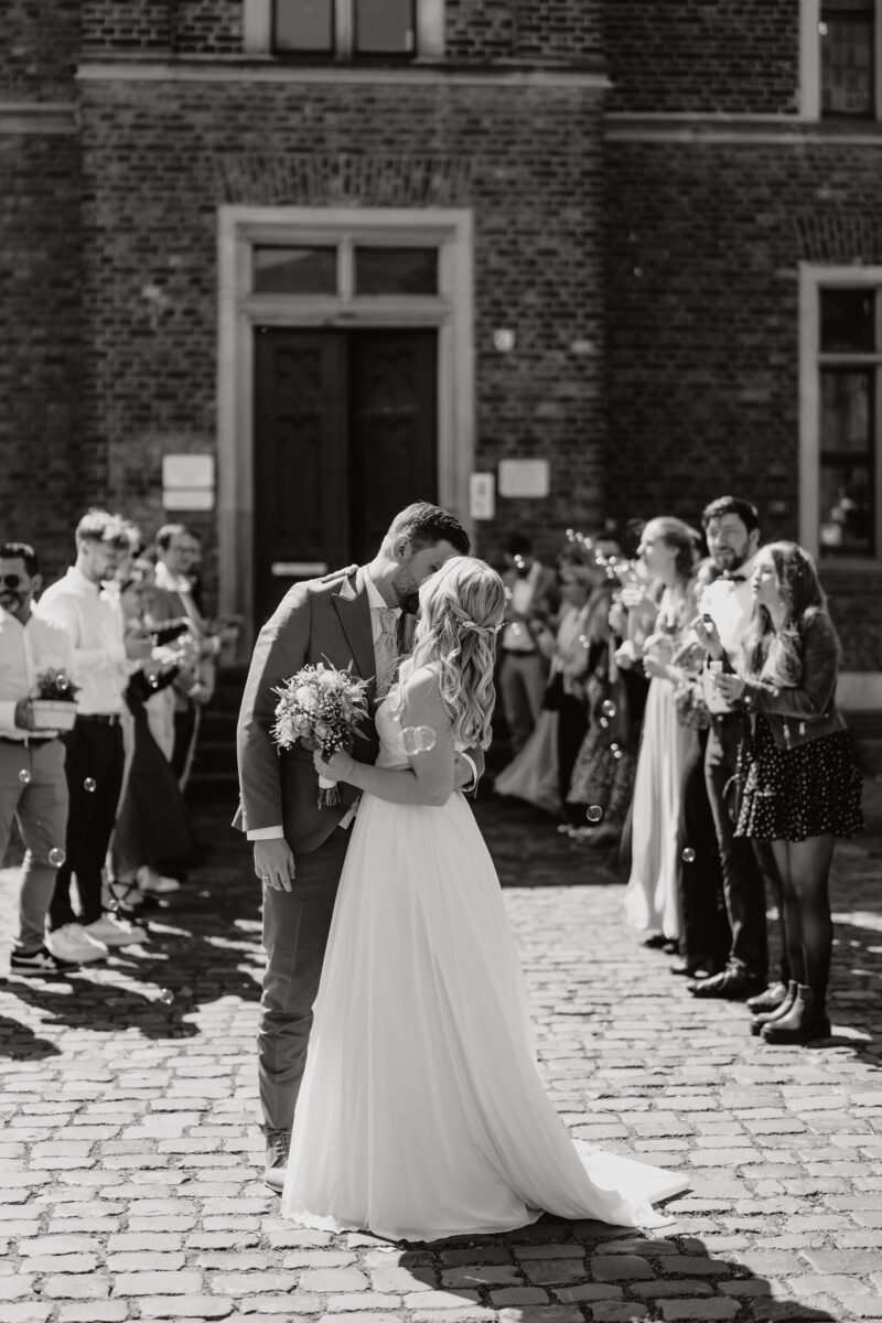 Hochzeitsportfolio, Braut und Bräutigam küssen sich. Um sie herum sind die Gäste in einem Sparlier und jubeln.