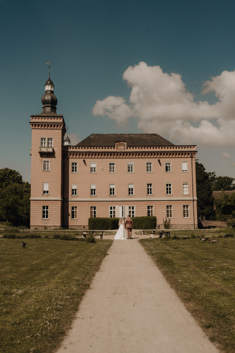 Hochzeitsportfolio, Totale, Braut und Bräutigam stehen vor einem Schloss, in der Mitte eines Weges. Si e halten sich in den Armen und küssen sich.