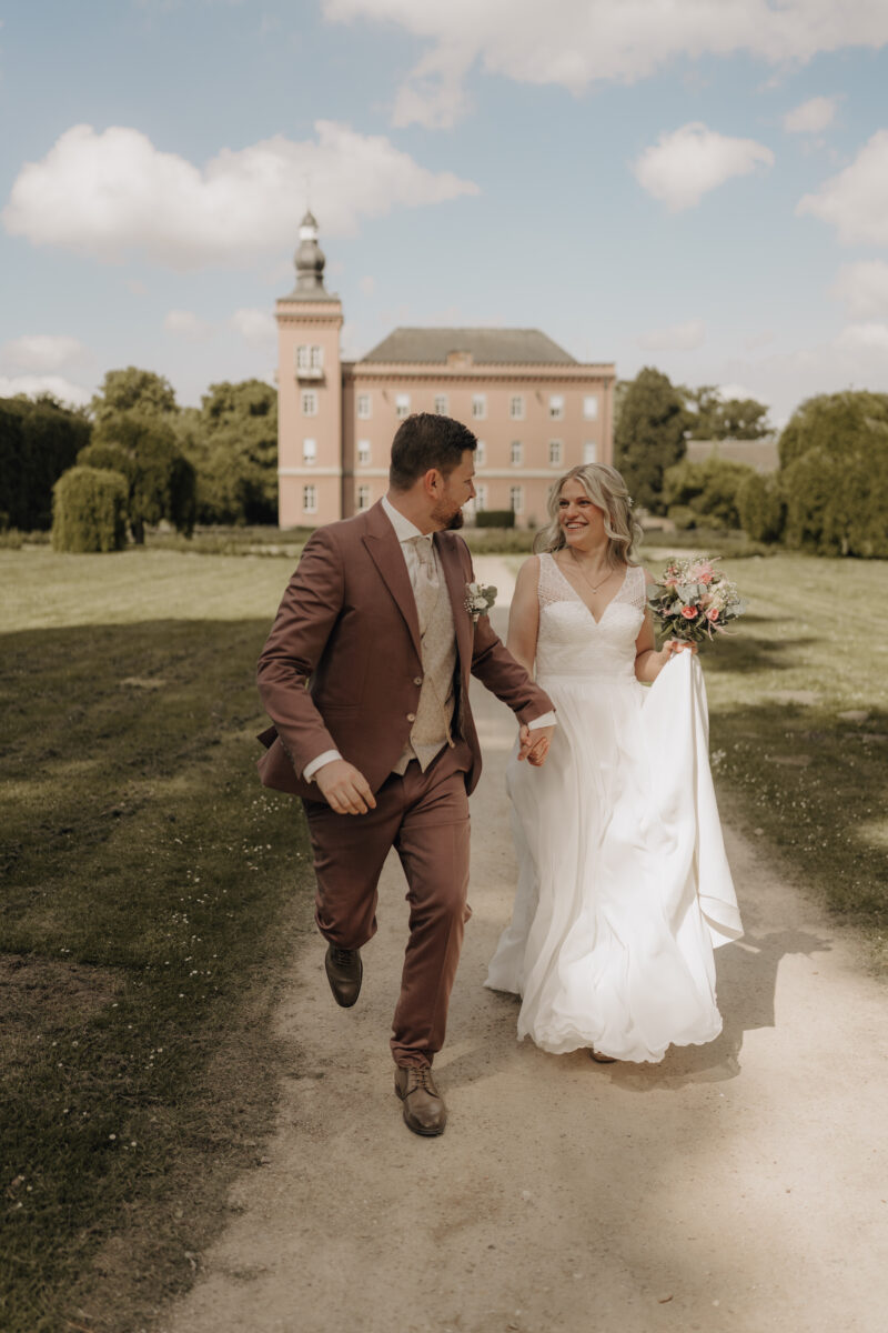 Hochzeitsportfolio, Brautpaar läuft gemeinsam vor einem Schloss durch den Garten auf die Kamera zu | Hochzeitsfotografie NRW