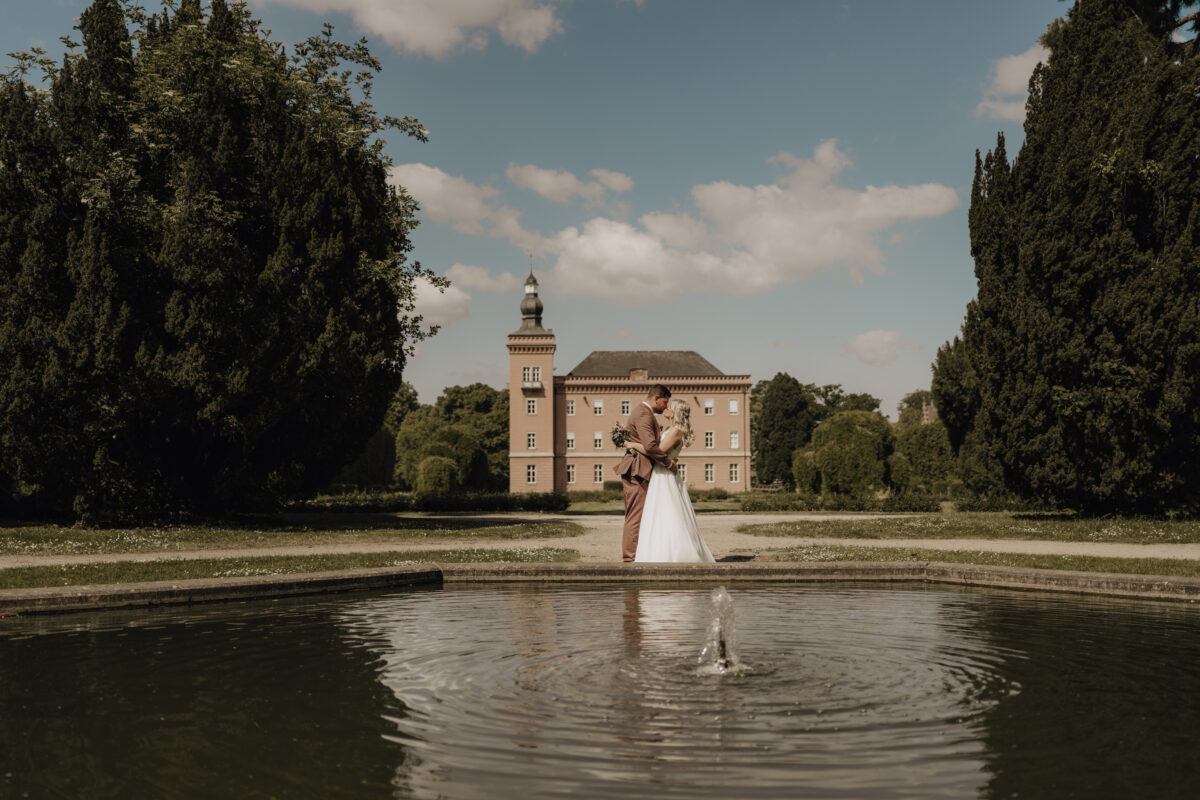 Hochzeitsportfolio, Braut und Bräutigam stehen in der Ferne vor einem Schloss. Im Vordergrund ist ein kleiner See. Beide halten sich in den Armen und lächeln sich verliebt an.