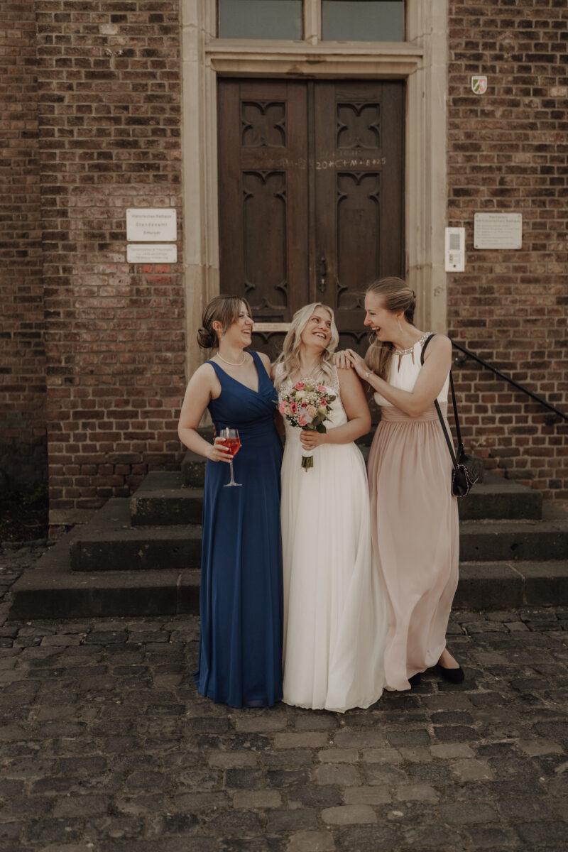 Hochzeitsportfolio, Brautbild mit zwei Freundinnen. Die Braut steht in der Mitte umgeben von ihren zwei Freundinnen. Freundinnen schauen die Braut an und lächeln. Die Braut lächelt.