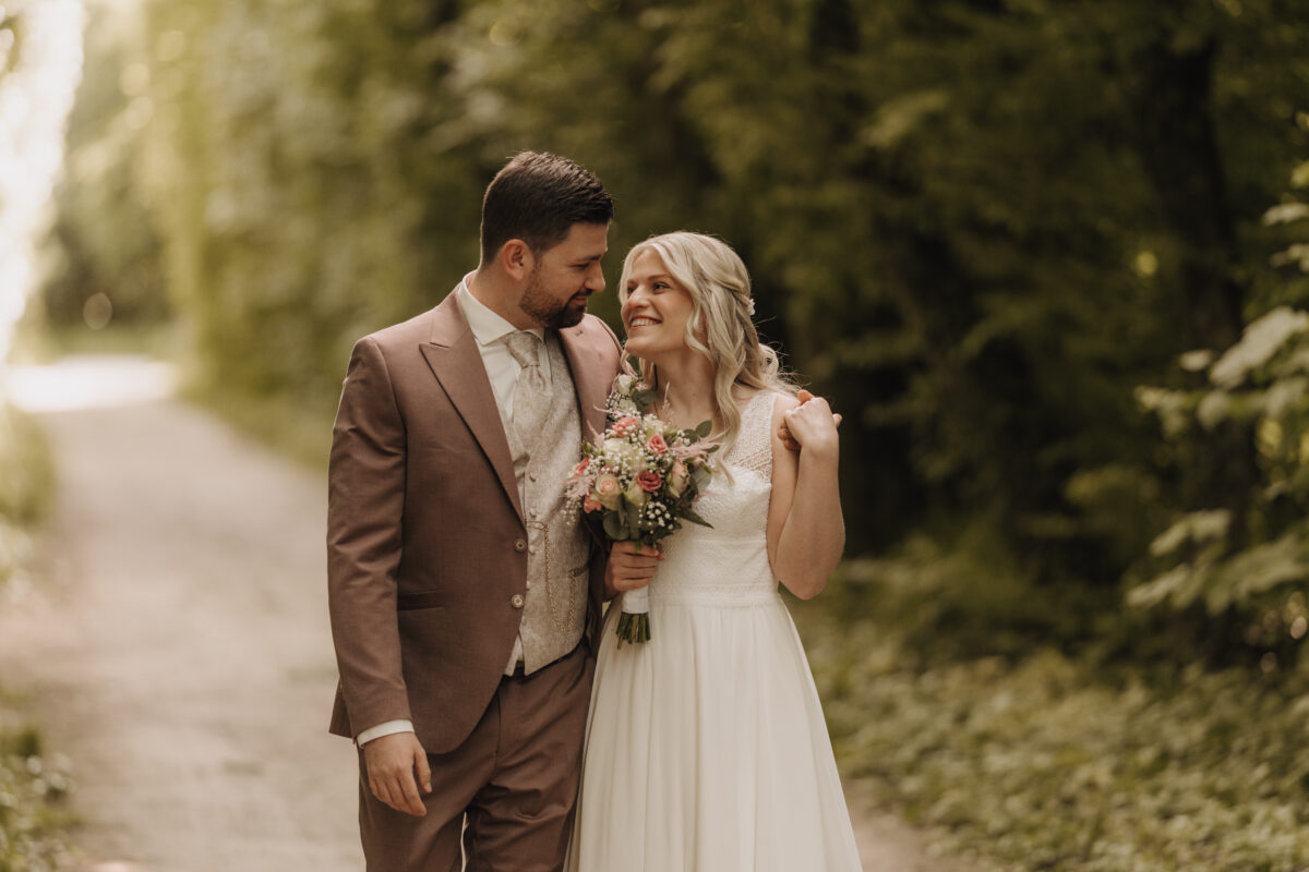 Hochzeitsportfolio, Braut und Bräutigam laufen einen Weg entlang. Er hat seinen Arm um ihre Schulter gelegt. Beide lächeln sich an.