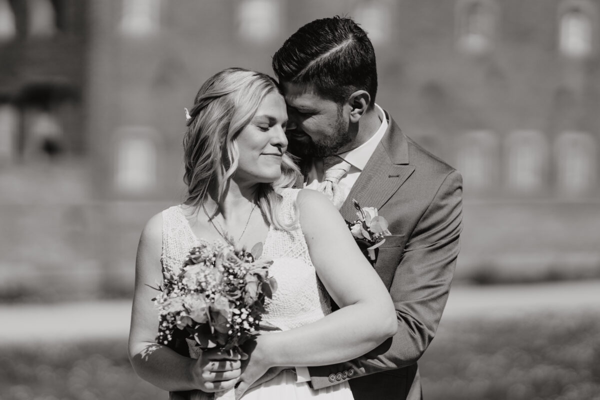 Hochzeitsportfolio, Bräutigam umarmt die Braut. Sein Gesicht ist ihrem ganz nah. Sie schaut lächelnd zur