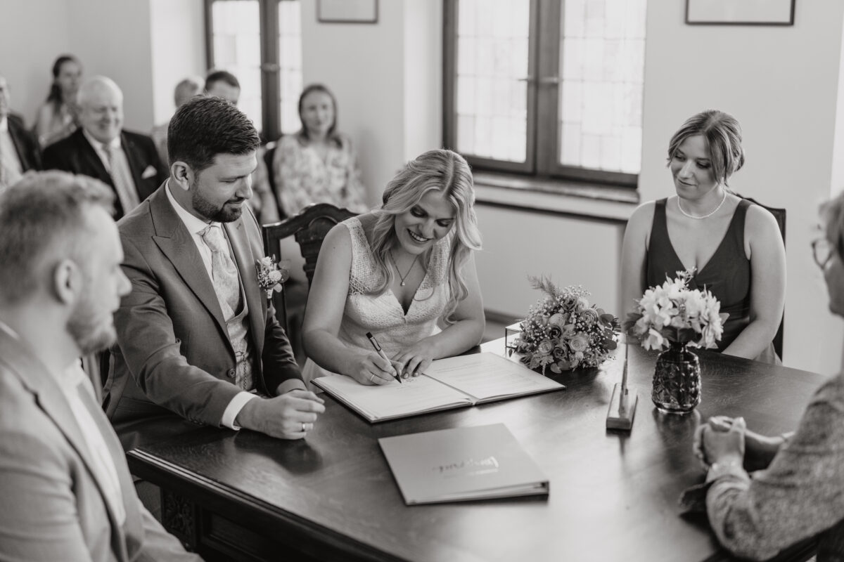 Hochzeitsportfolio, Braut und Bräutigam beim Standesamt. Braut unterschreibt die Eheurkunde. Beide lächeln.