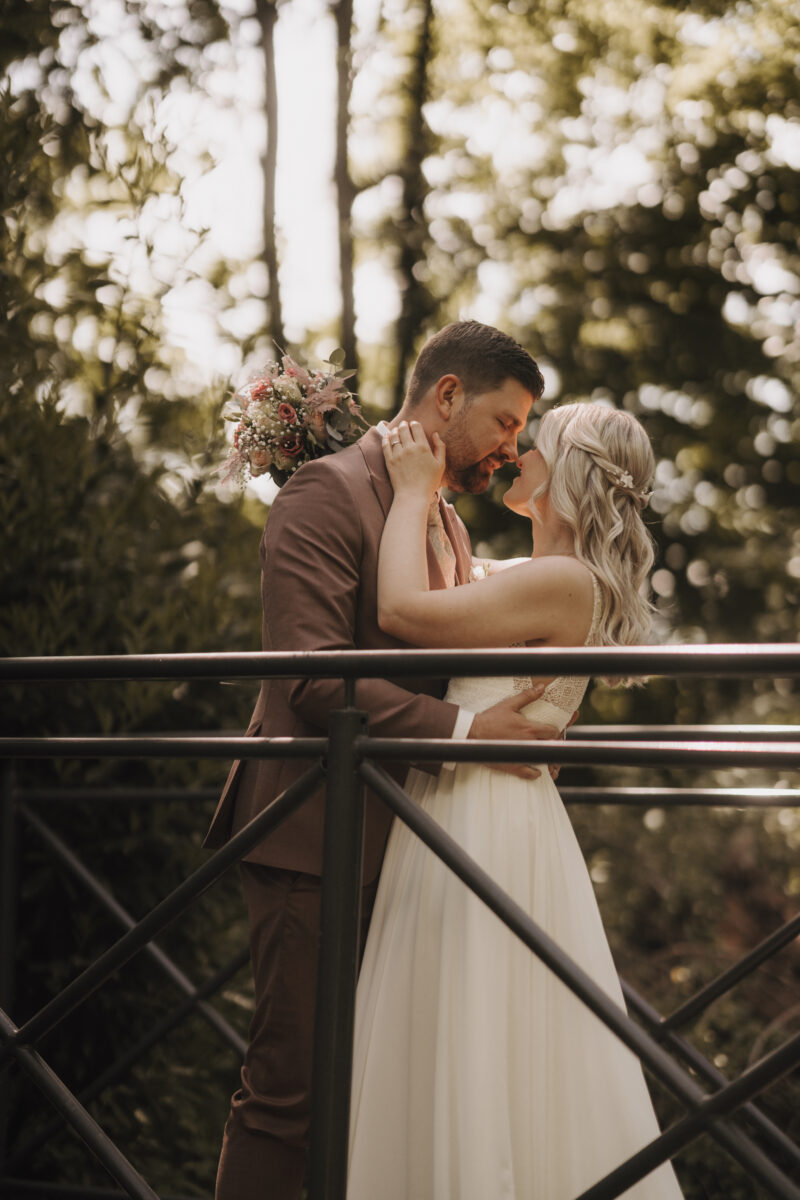 Hochzeitsportfolio, Braut und Bräutigam stehen auf einer Brücke. Seine Hände liegen auf ihrer Taille. Ihre Hände liegen auf seinen Schultern. Ihre Nasenspitzen berühren sich.