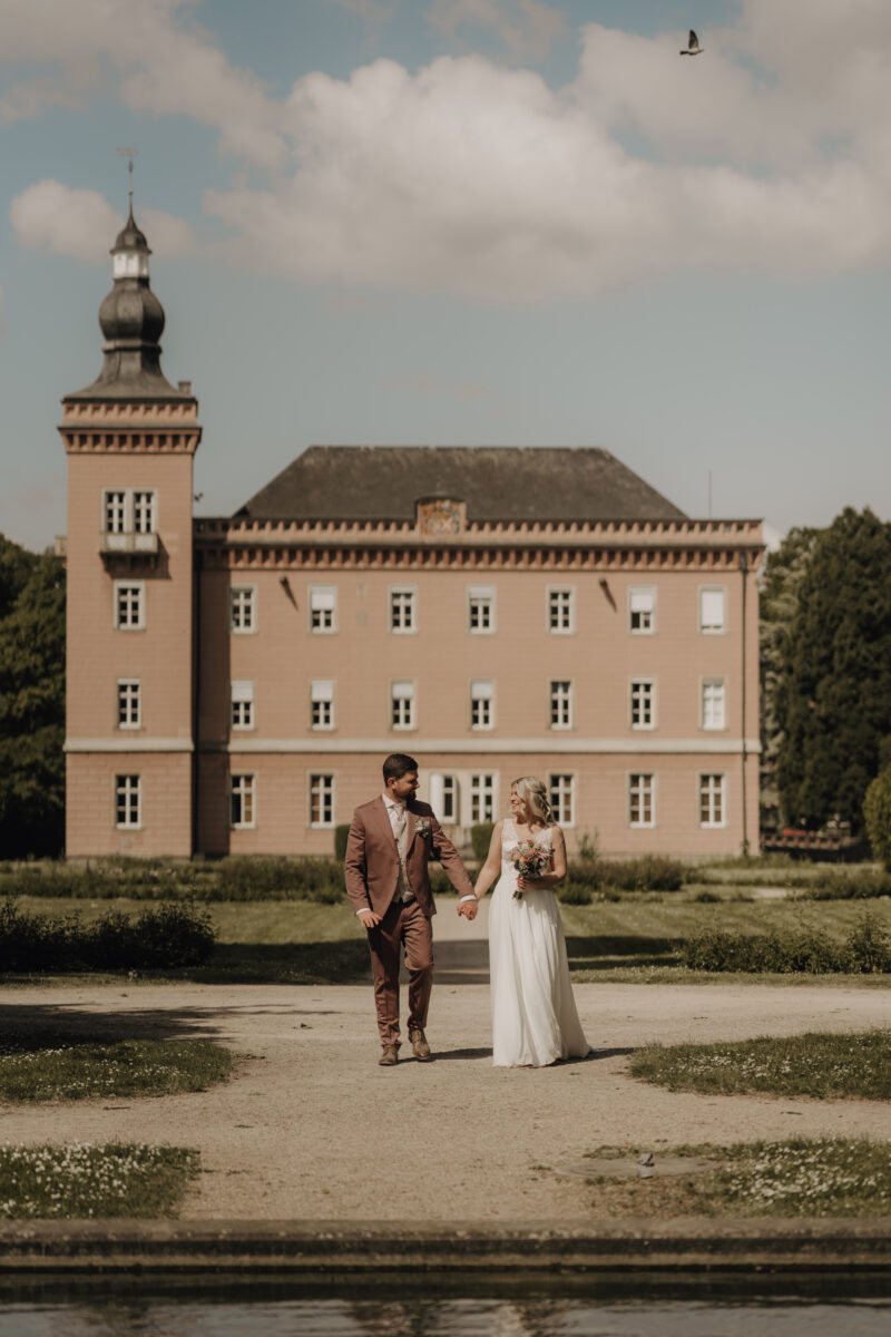 Hochzeitsportfolio, Brautpaar vor einem historischen Schloss und läuft auf die Kamera zu, sie gucken sich währenddessen tief in die Augen