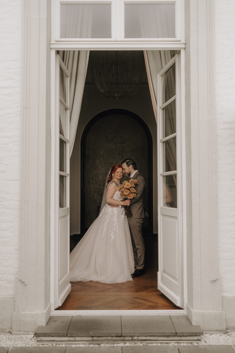 Hochzeitsportfolio, Brautpaar steht gegenüber von einander in einer eleganten Türöffnung eines Schlosses, Bräutigam lehnt seinen Kopf an den Kopf der Braut und hat dabei die Augen geschlossen | Hochzeitsfotografin NRW