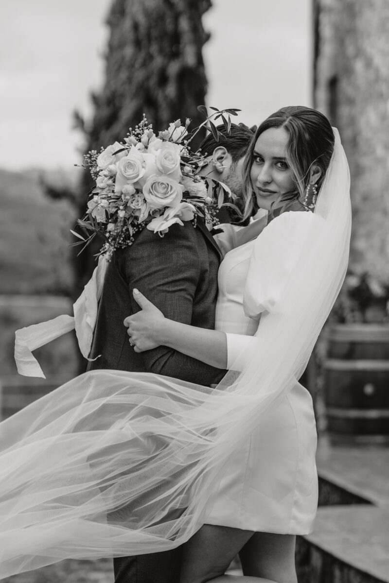 Hochzeitsportfolio, Braut und Bräutigam Porträit. Braut schaut in die Kamera und hält sich an seinem Oberarm fest. Bräutigam flüstert ihr etwas in das Ohr.