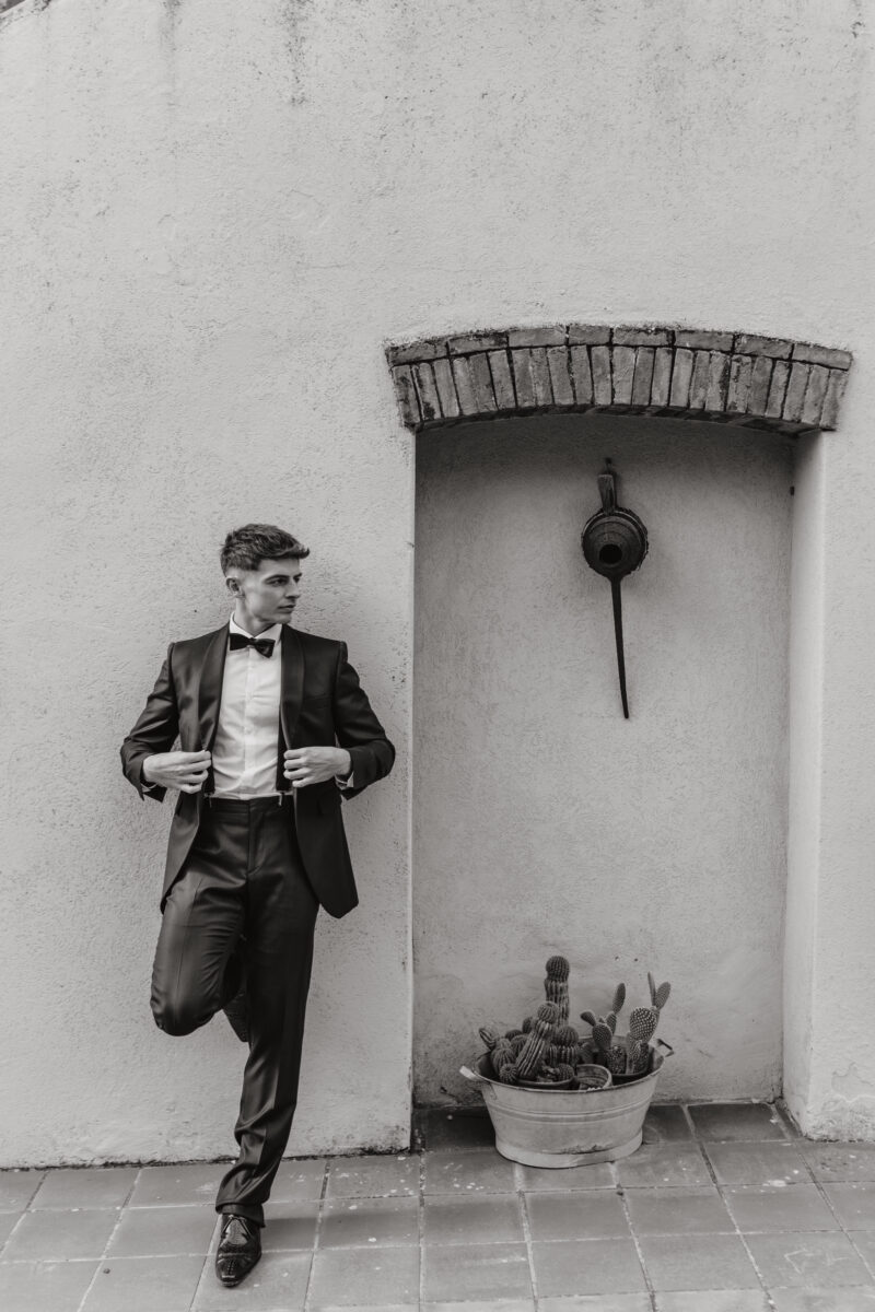Hochzeitsportfolio, Bräutigam steht angelehnt an der Wand und ist dabei sich das Sakko zu zumachen und guckt dabei nach links in die Ferne