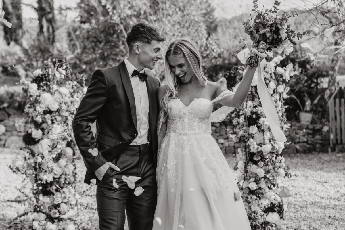 Hochzeitsportfolio, Braut und Bräutigam feiern draußen. Beide stehen nah aneinander. Sie hat ihren Arm mit dem Brautstrauß in die Luft gereckt. Er schaut sie lächelnd an.